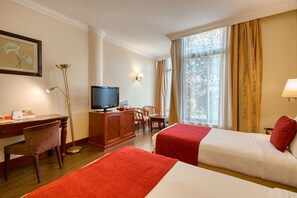 Habitación de hotel con dos camas, un escritorio, un televisor y un ventanal grande con cortinas.