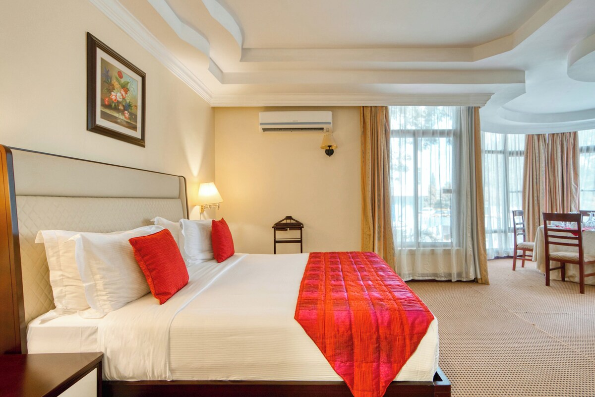 A hotel room with a large bed, a bedside table, a lamp, an air conditioning unit, a window with curtains, and a chair.