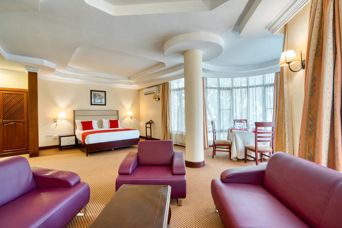 A hotel room with a bed, two purple sofas, a small table, and a large window with curtains.