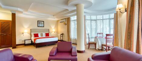Quarto de hotel com cama, dois sofás roxos, uma mesinha e uma janela grande com cortinas.