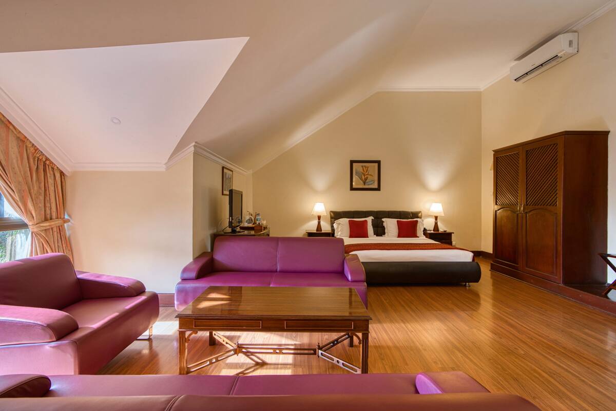 A hotel room with a wooden floor, a purple sofa, a wooden coffee table, a bed with red and white bedding, and a wardrobe.