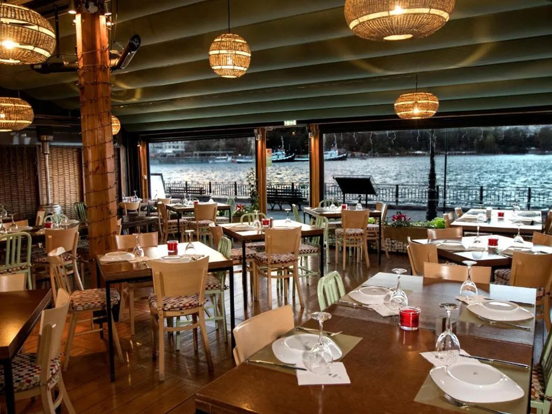 Een restaurant met gedekte tafels, grote ramen met uitzicht op een wateroppervlak en hangende rieten lampen.