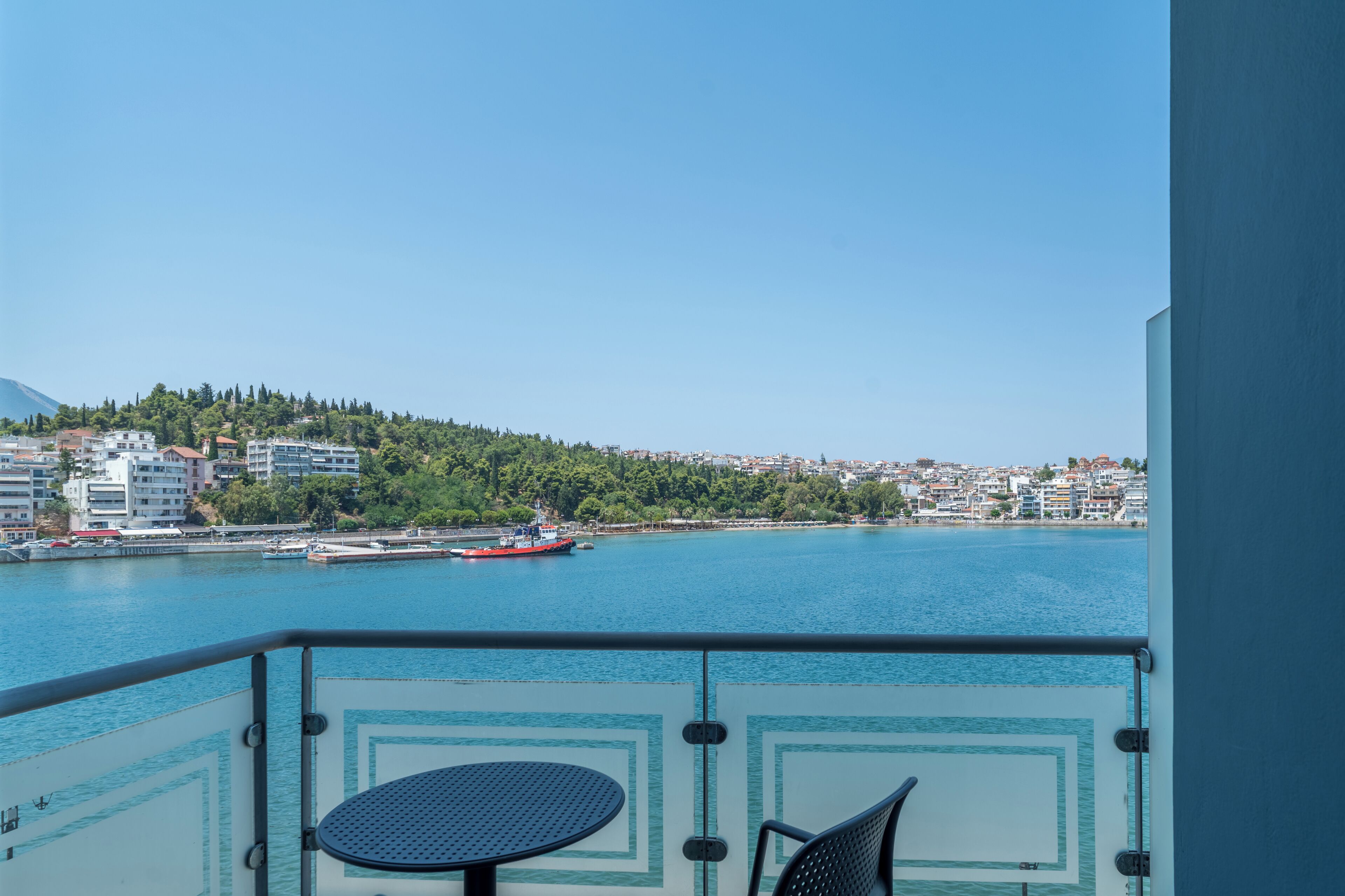 Un balcón con mesa y silla, con vista a una masa de agua donde se ve un barco rojo y un paisaje urbano al fondo.