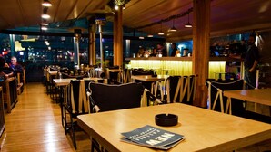A restaurant with wooden tables and chairs, a bar area, and a view of the city at night. - Paliria Hotel (Chalcis)