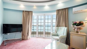 A living room with a TV on a stand, a chair, a small table, and a view of the sea through large windows. - Paliria Hotel (Chalcis)