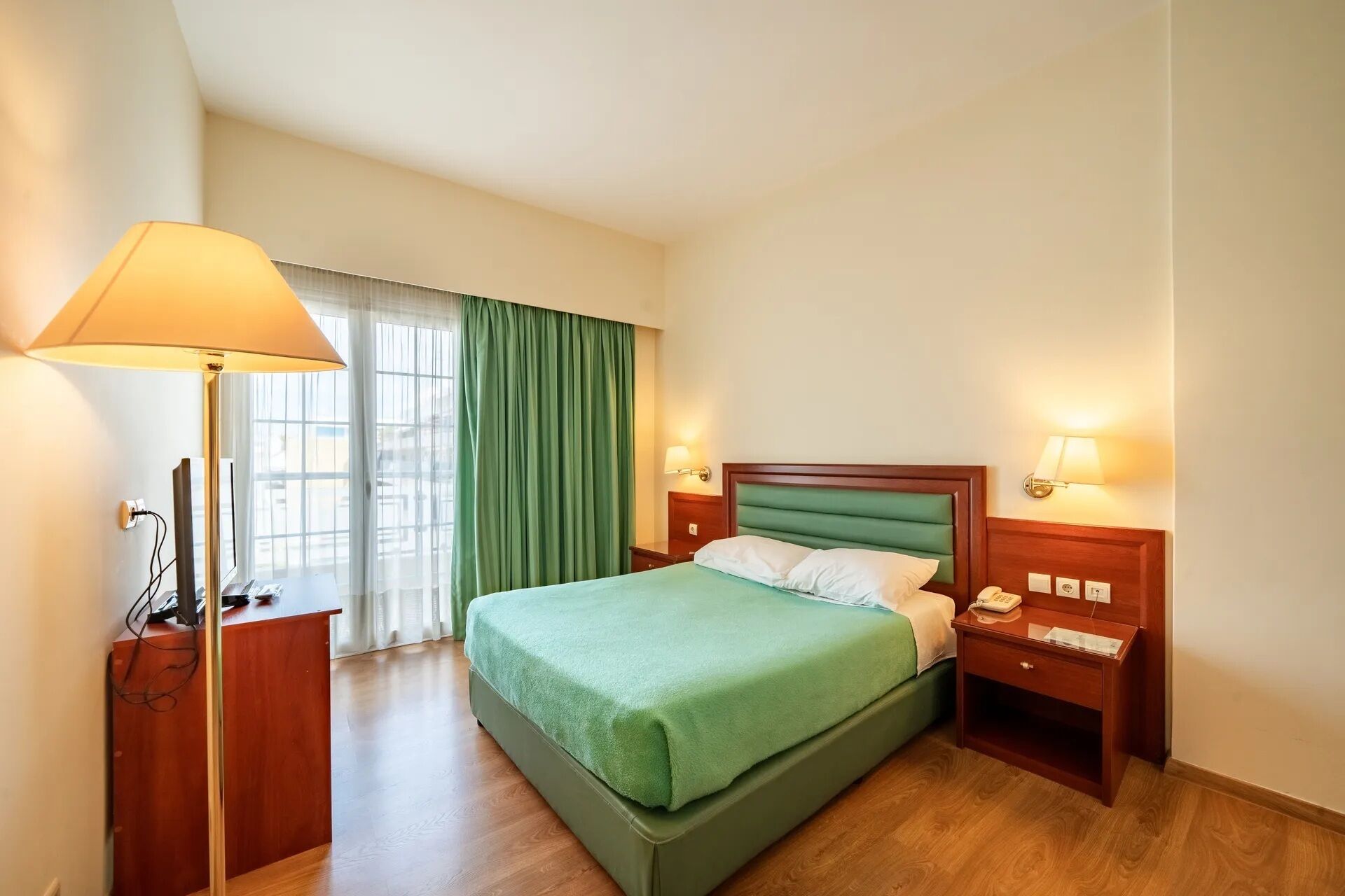 Una habitació d'hotel amb un llit, làmpades de nit, un escriptori amb un telèfon i una finestra amb cortines.