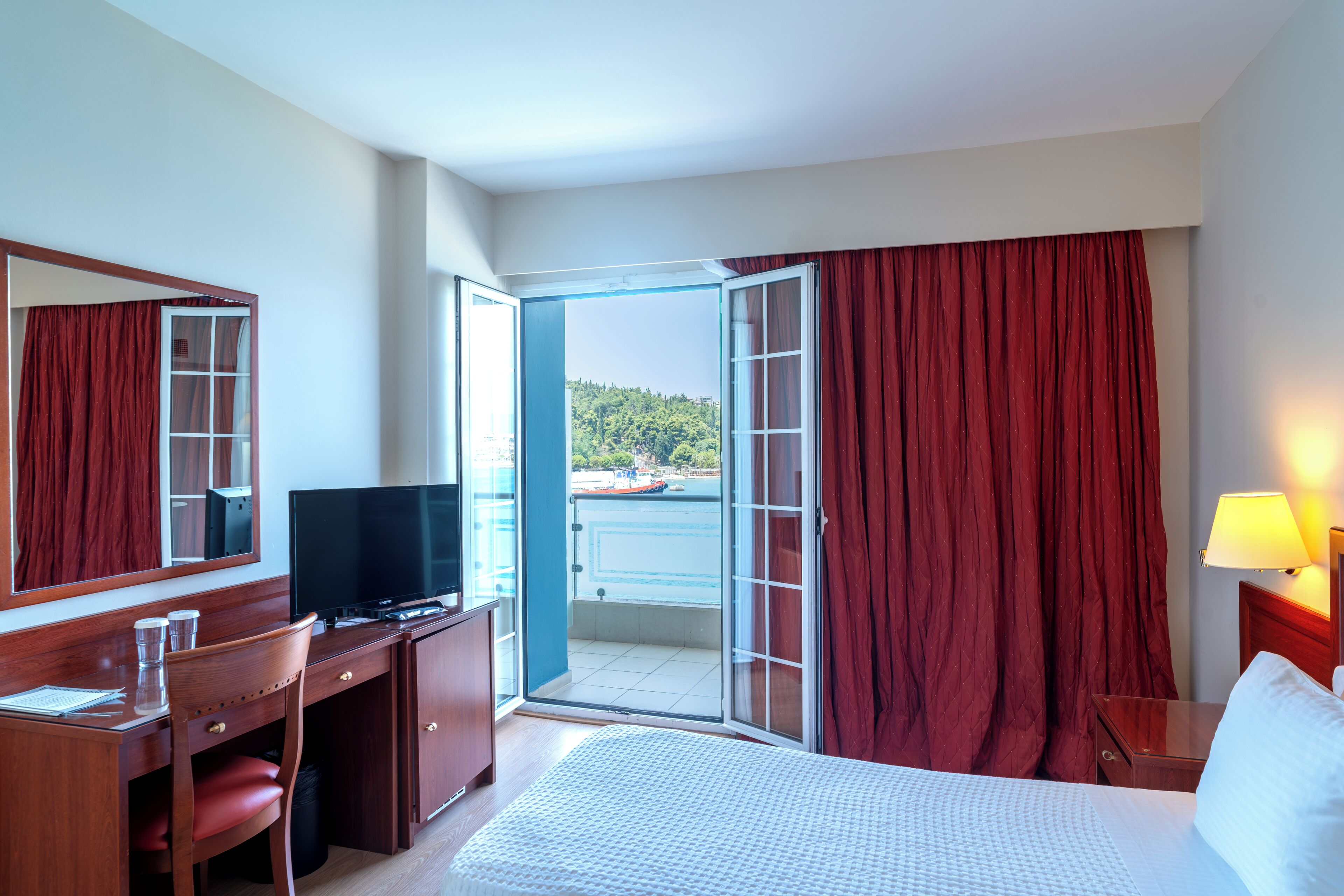 Una camera d'albergo con un letto, una scrivania con televisore, una sedia, una lampada e vista su un corpo d'acqua attraverso una porta aperta.