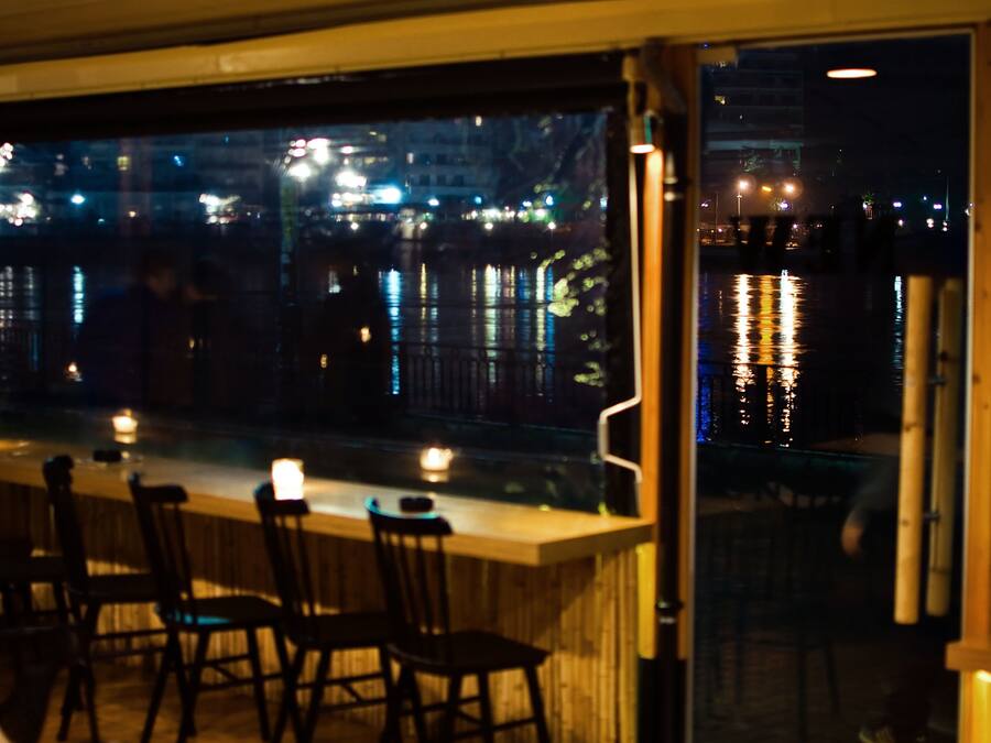 Zona de bar con sillas y mesas, vista nocturna de un paisaje urbano y un cuerpo de agua que refleja luces.