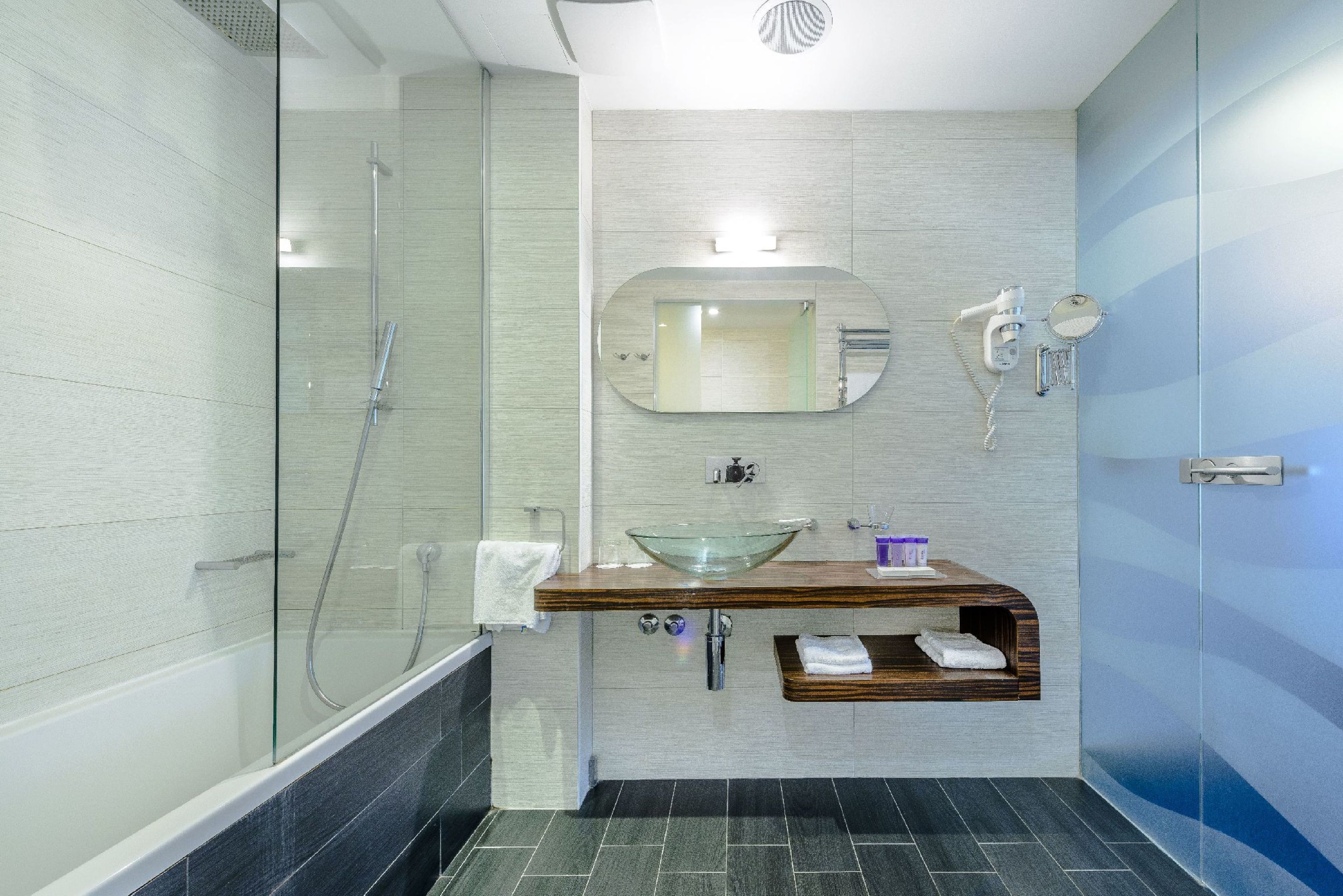 Un baño moderno con ducha envuelta en vidrio, tocador de madera con lavabo de cristal y espejo sobre el lavabo.