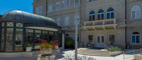 A historic building with a sign reading "Hotel Lapad." It features a covered entrance, a fountain, and a well-maintained garden area.