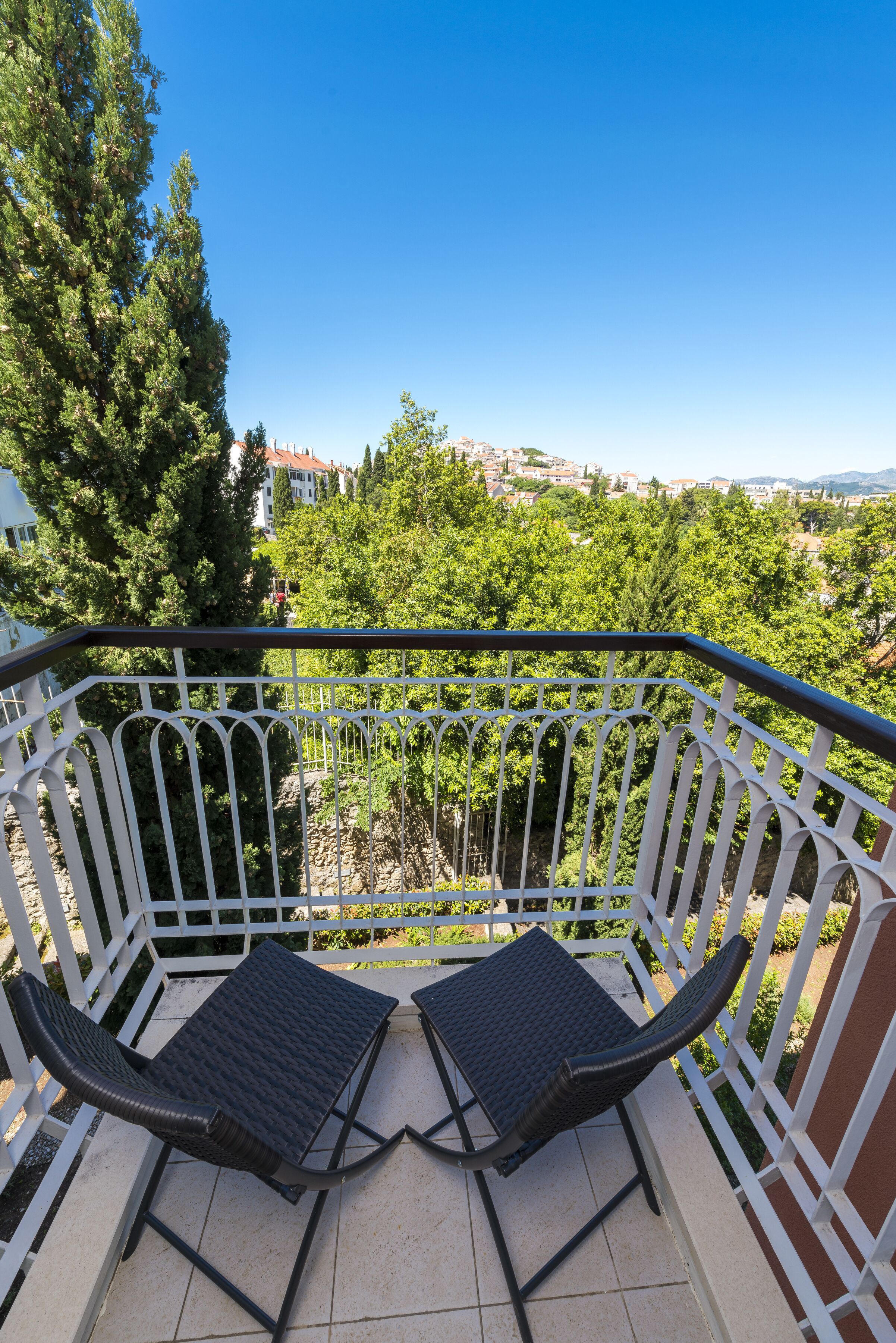 A balcony with two black chairs and a white railing, overlooking a green landscape and buildings.