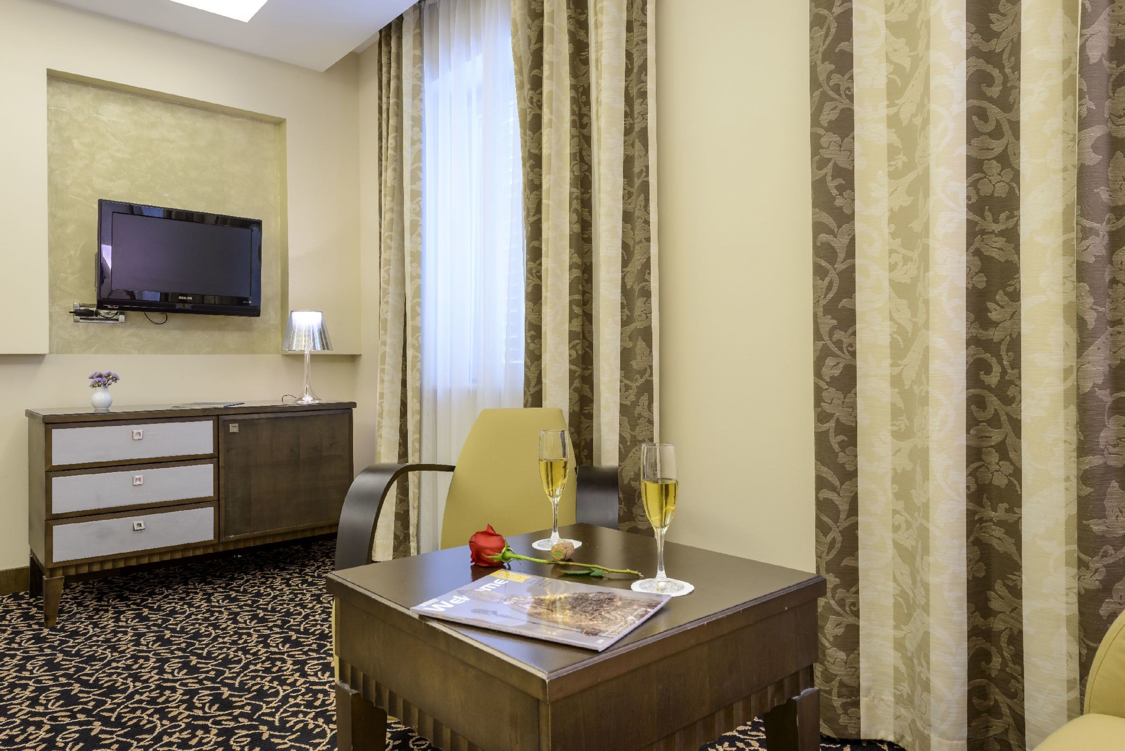 Hotelkamer met een flatscreen televisie aan de muur, een houten bureau met een tijdschrift, twee champagneglazen en een roos op de tafel.