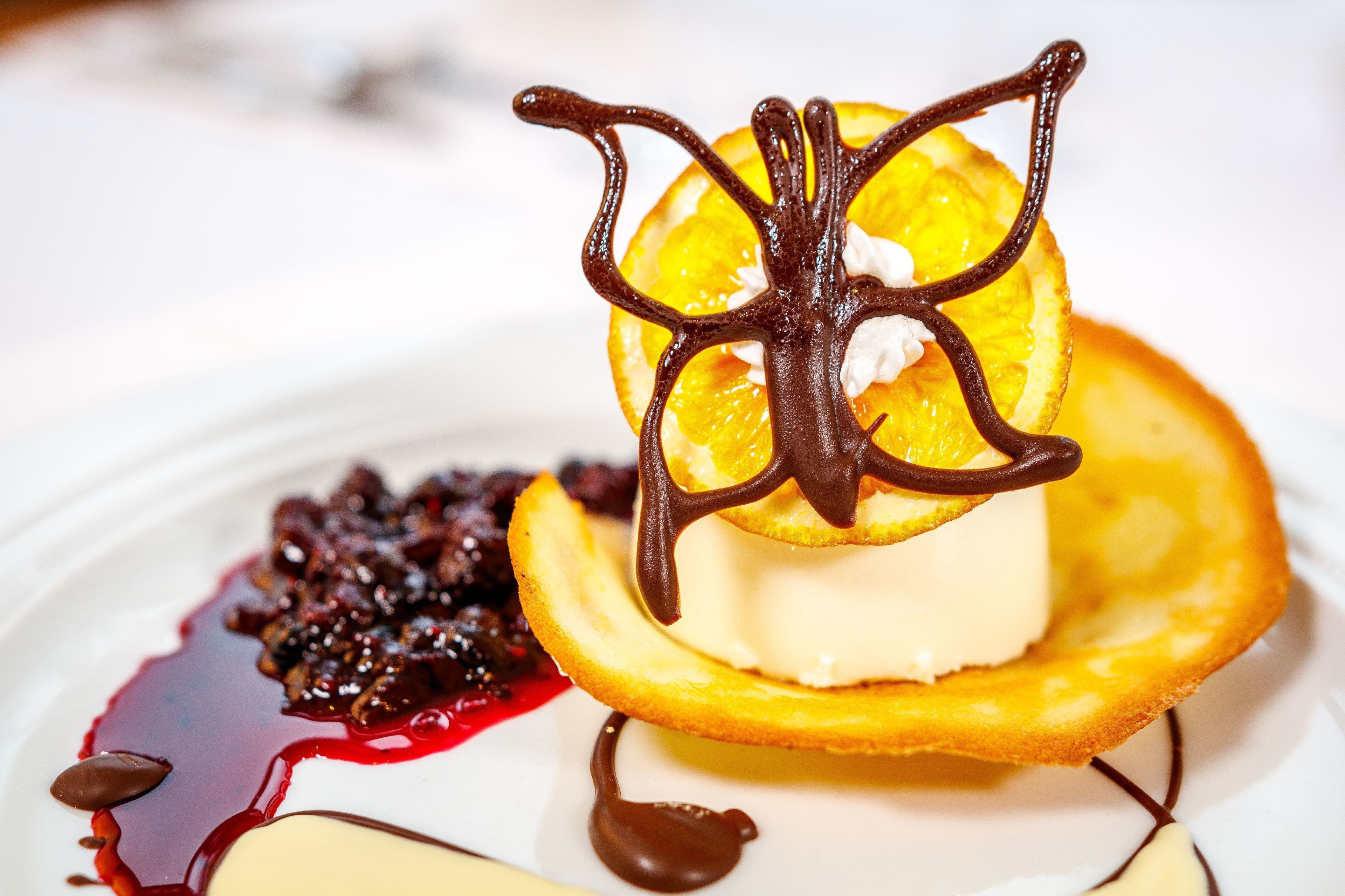 Un postre con telaraña de chocolate sobre una rodaja de naranja, rodeado de una base cremosa y un puré de frutos rojos.