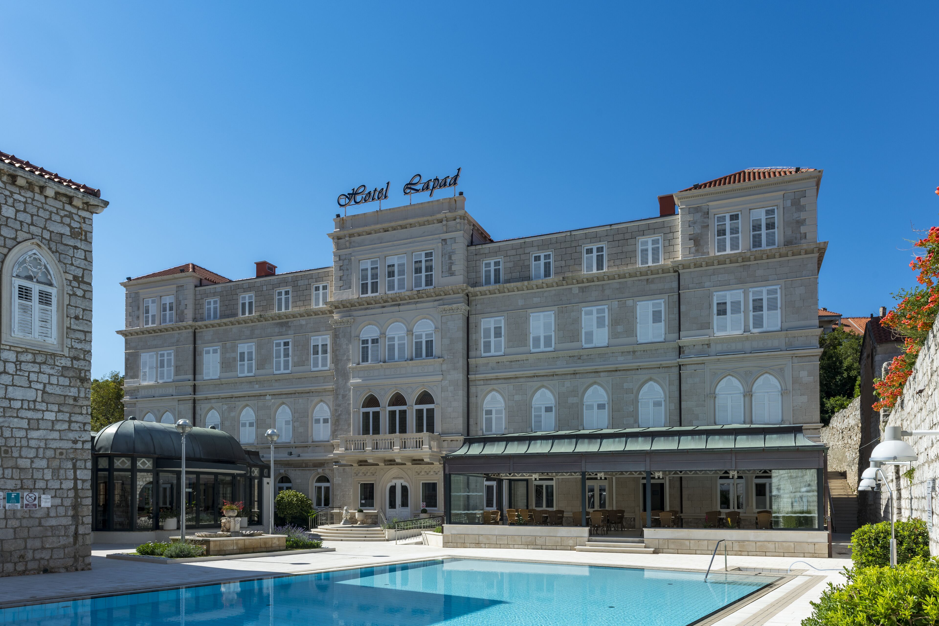 Ein großes, historisches Gebäude mit einem Schild, auf dem "Hotel Lapad" steht. Es hat mehrere Stockwerke mit Rundbogenfenstern und im Vordergrund einen Swimmingpool.