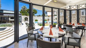 A restaurant with outdoor seating, large windows, and a view of a pool and buildings. - Hotel Lapad (Dubrovnik)