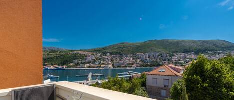 A balcony with a table set for two, overlooking a coastal town with a clear view of the sea and hills.