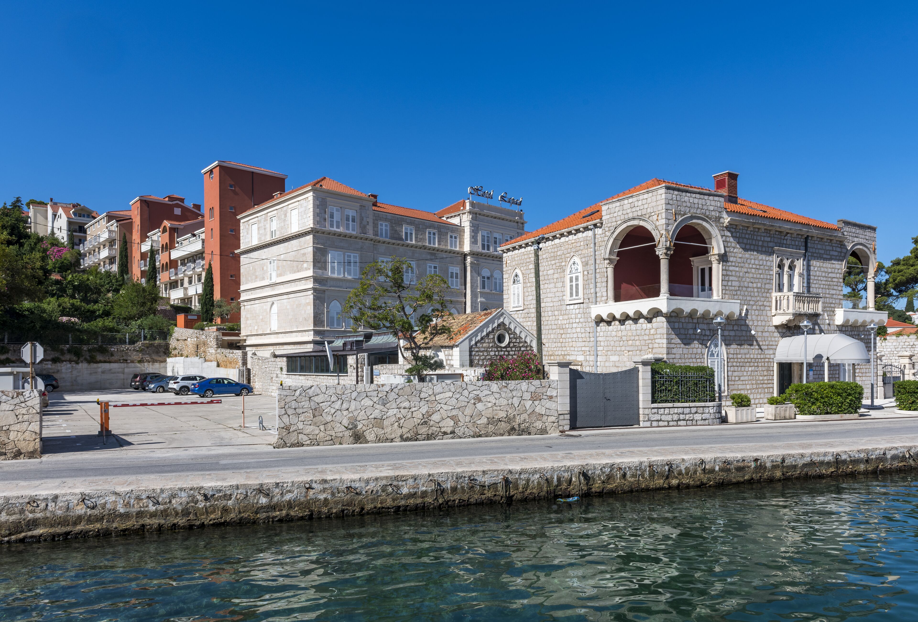 Un hotel sul lungomare con architettura in pietra, vista chiara sull'acqua e un'area di parcheggio con auto.