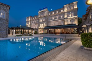 Hotel features a large rectangular pool surrounded by a paved area with potted plants, set against a backdrop of its classical architecture.