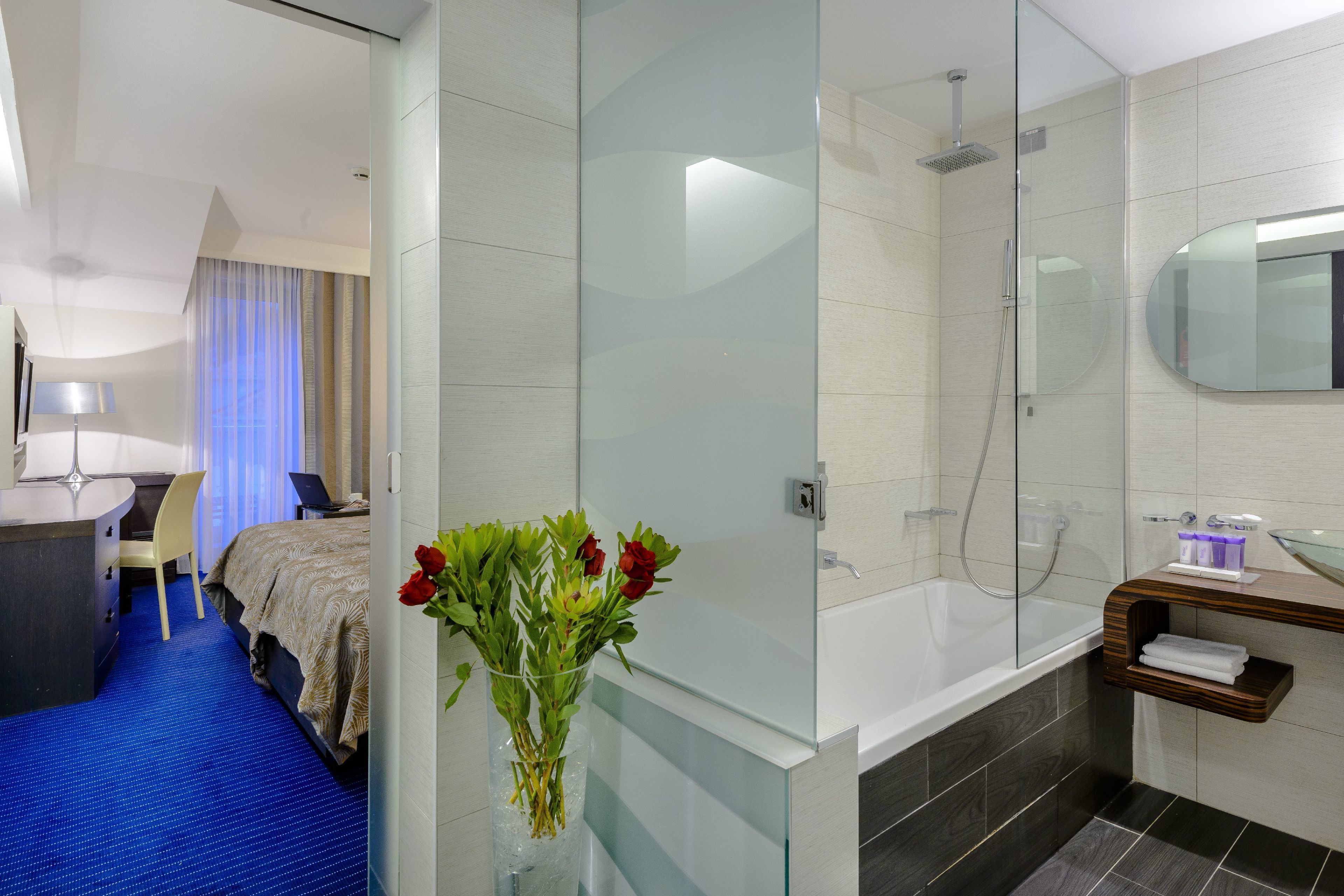 A modern hotel room with a bed, a desk, a laptop, a vase with flowers, a shower, and a glass-enclosed bathroom.