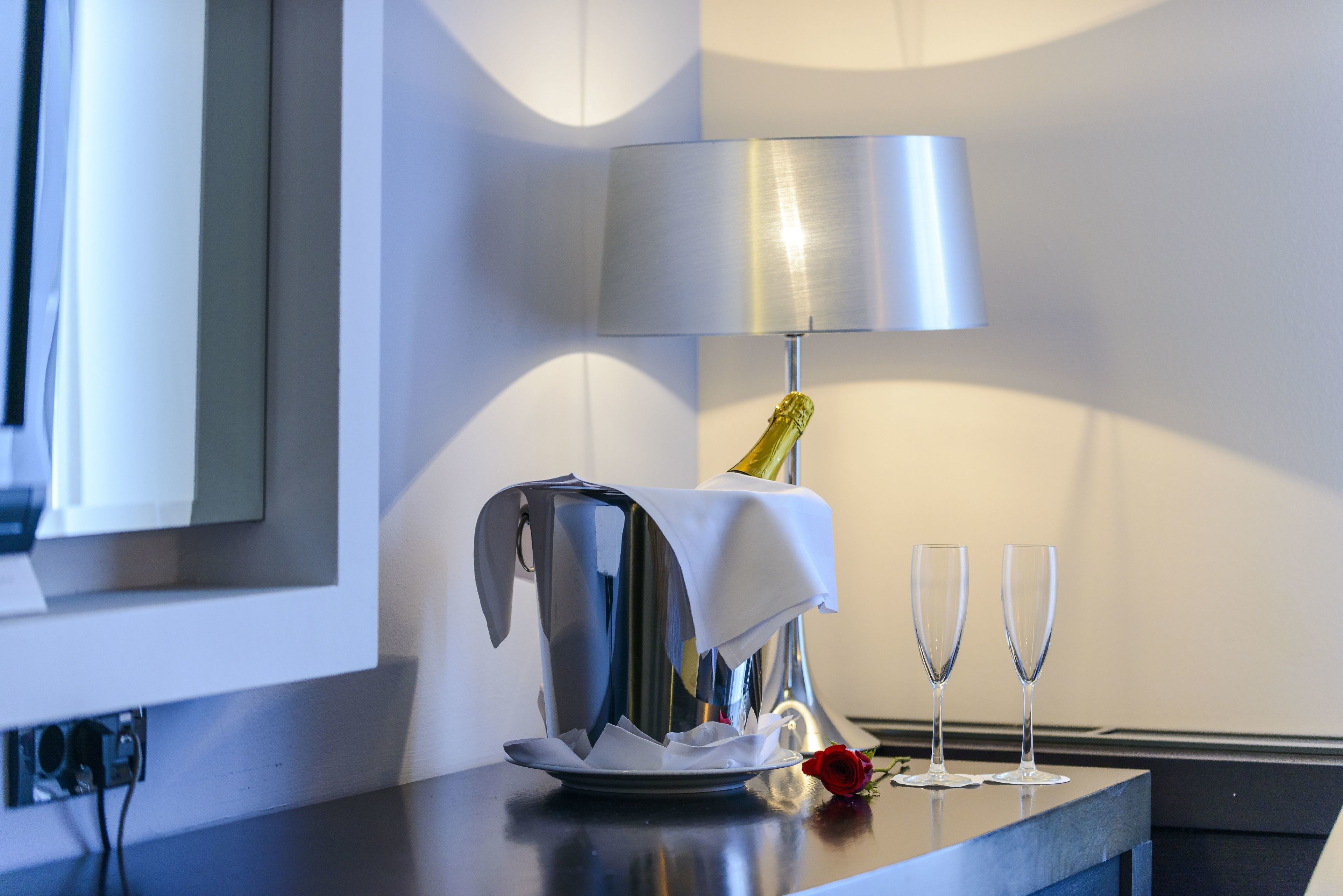 Habitación de hotel con una mesa metálica, una botella de champán, dos copas y una rosa roja.