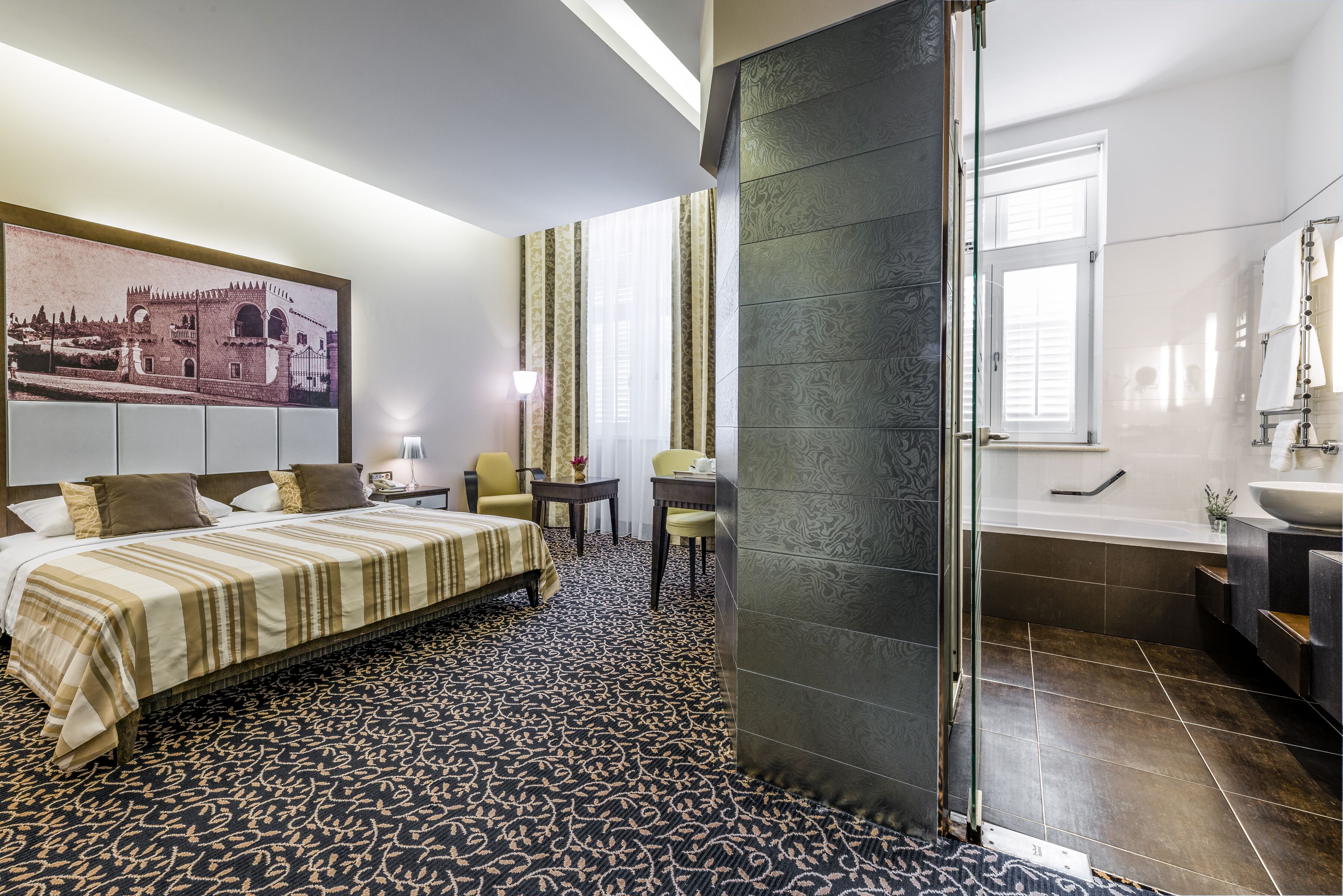 Ein Hotelzimmer mit Bett, Schreibtisch, Stuhl sowie einem Badezimmer mit Badewanne und einem großen Fenster.
