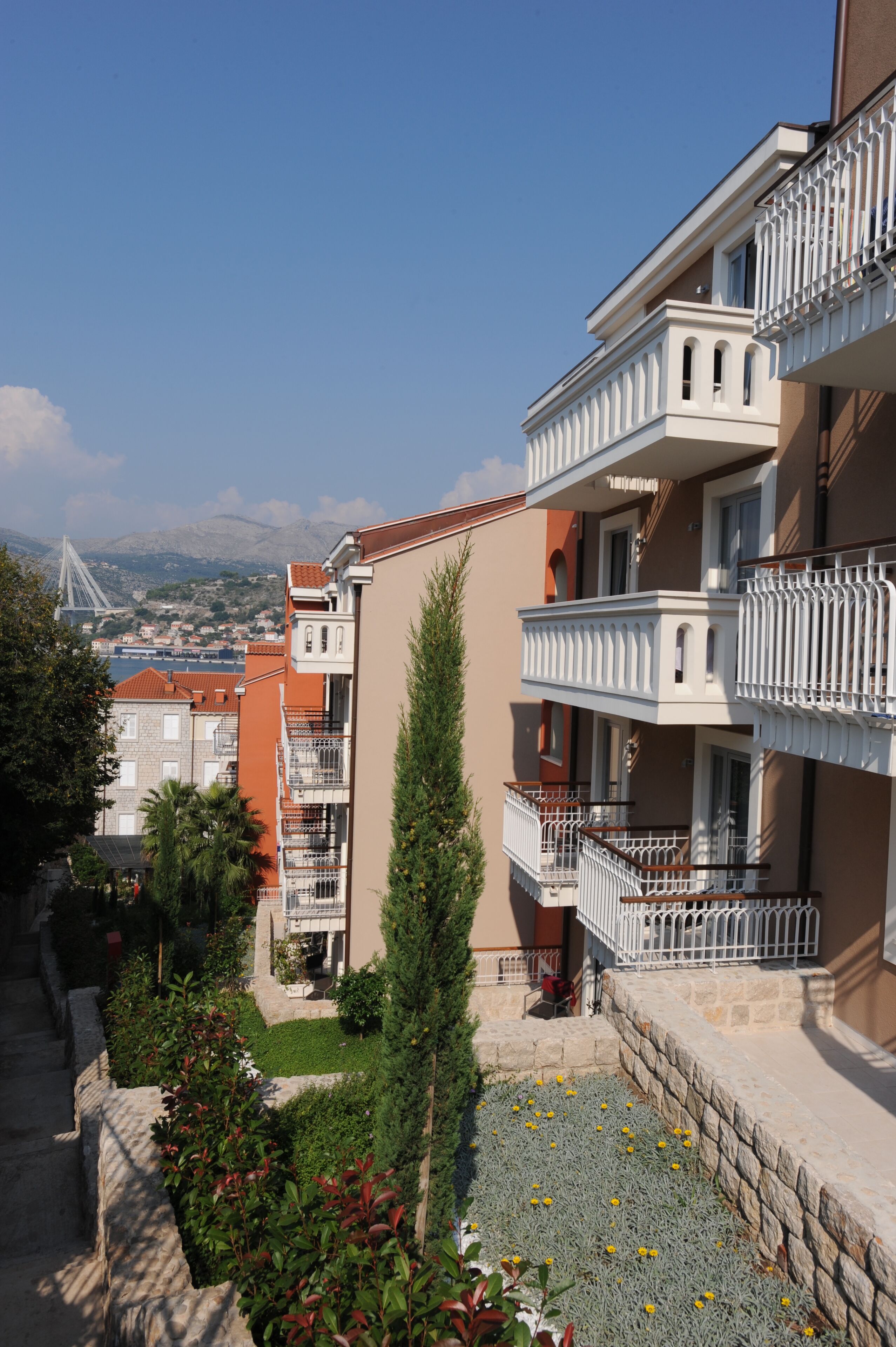 A multi-story building with balconies, a view of a bridge, and a courtyard with plants and flowers.