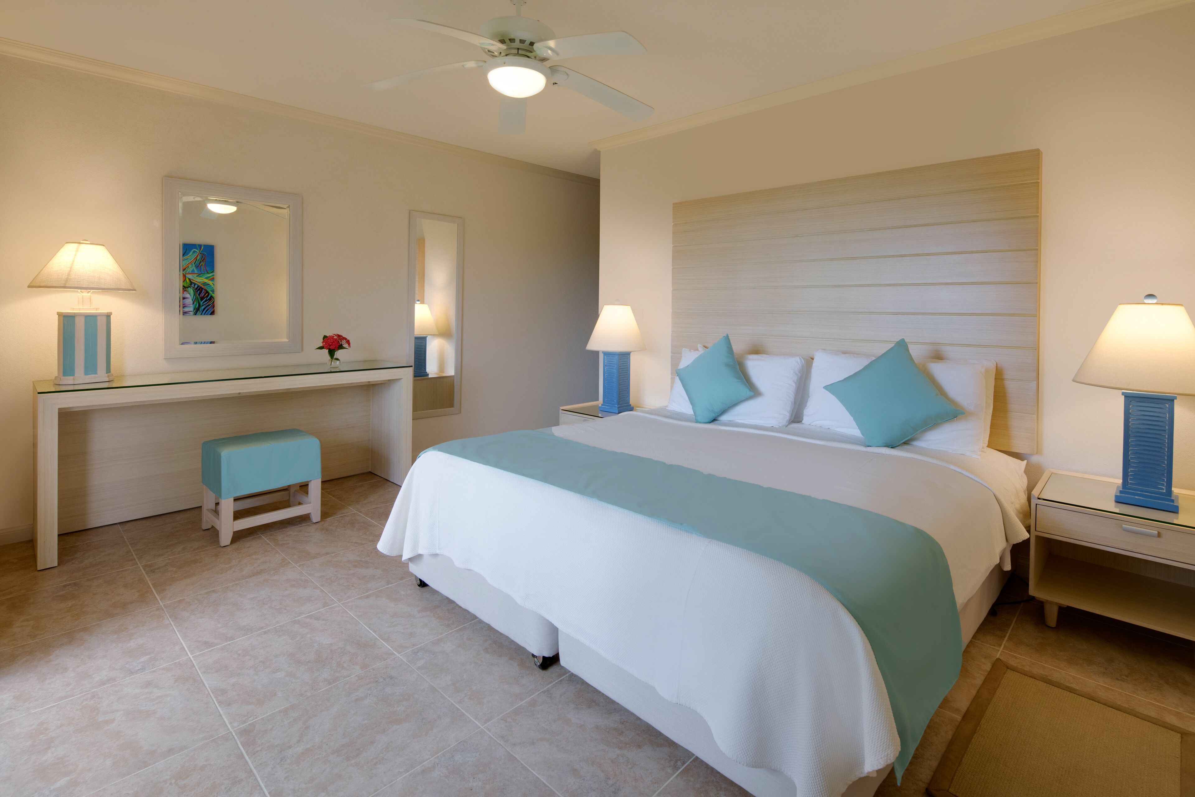 A hotel room with a large bed, a desk with a chair, a small stool, and two bedside tables with lamps.