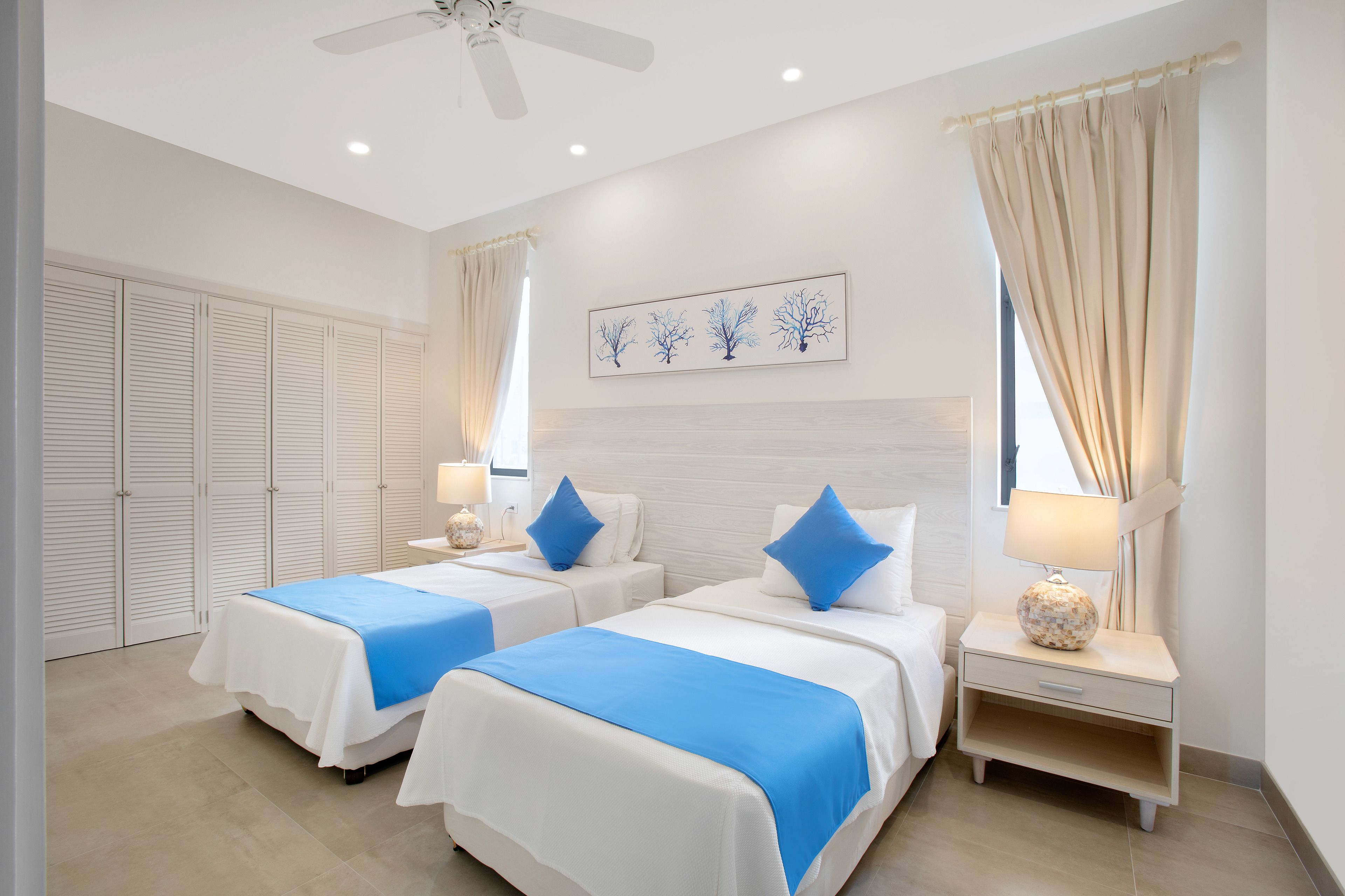 A hotel room with two beds, a ceiling fan, a nightstand with a lamp, and a window with curtains.