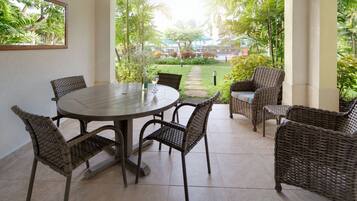 Une terrasse couverte avec une table ronde et des chaises en osier, entourée d’une végétation luxuriante et offrant une vue sur une piscine et des arbres.