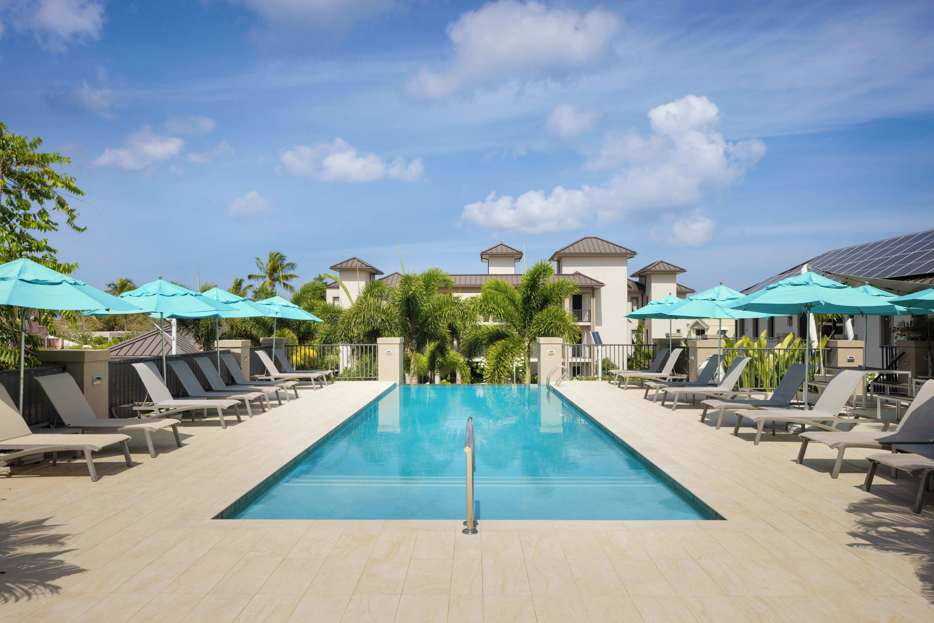 A swimming pool with lounge chairs and umbrellas, surrounded by buildings and palm trees.