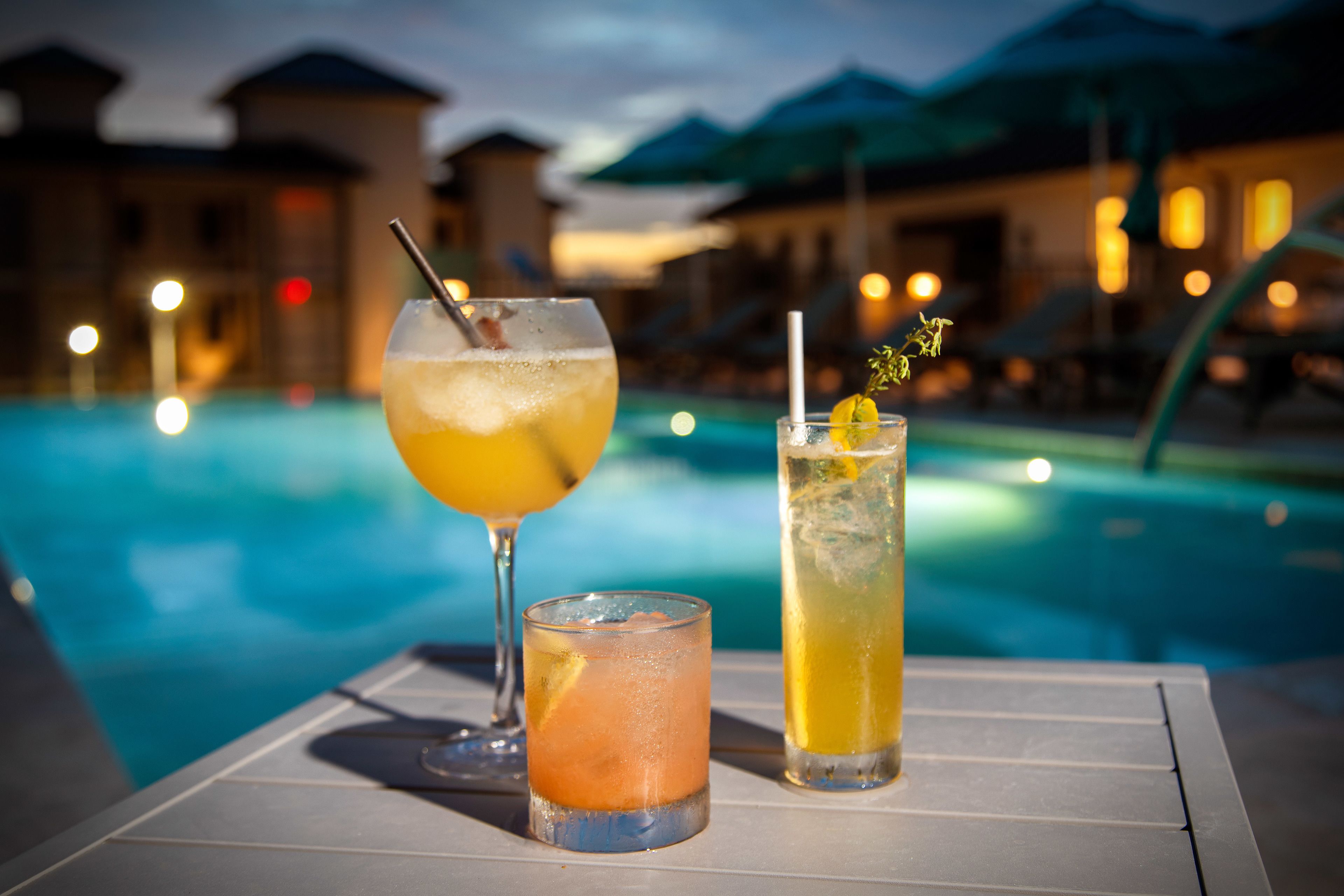 Três bebidas sobre uma mesa perto de uma piscina, com um edifício ao fundo.