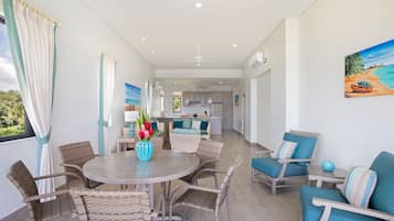 A modern kitchen with white cabinets and a central island, a dining area with wicker chairs and a round table, and a living area with blue upholstered chairs and a painting of a beach scene.