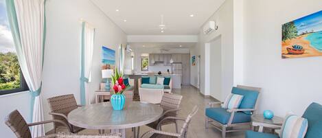 A modern kitchen with white cabinets and a central island, a dining area with wicker chairs and a round table, and a living area with blue upholstered chairs and a painting of a beach scene.