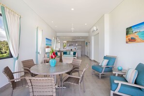 A modern kitchen with white cabinets and a central island, a dining area with wicker chairs and a round table, and a living area with blue upholstered chairs and a painting of a beach scene.