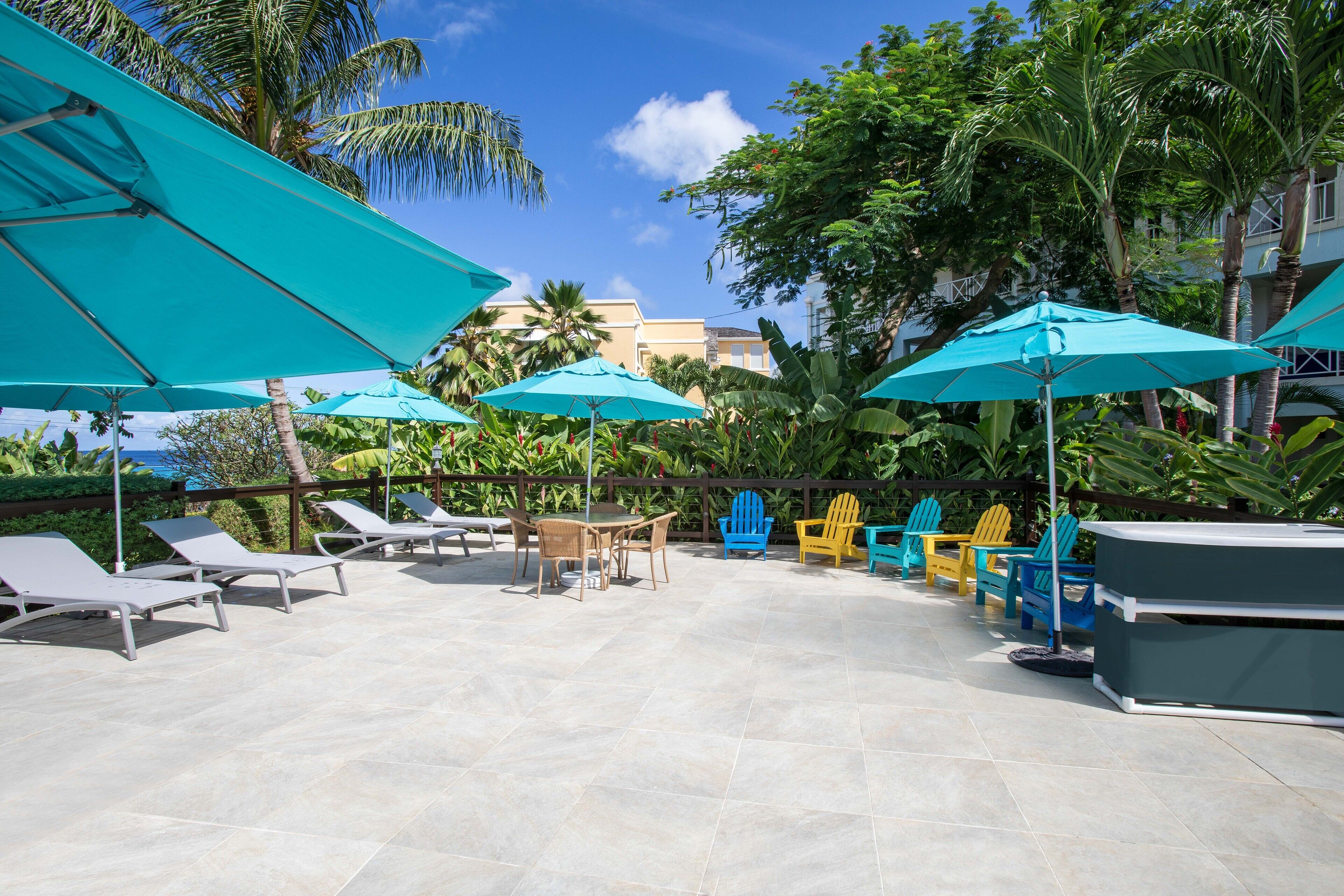 A patio area with lounge chairs, tables, and umbrellas, surrounded by lush greenery and palm trees.