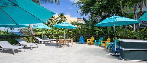 A patio area with lounge chairs, tables, and umbrellas, surrounded by lush greenery and palm trees.