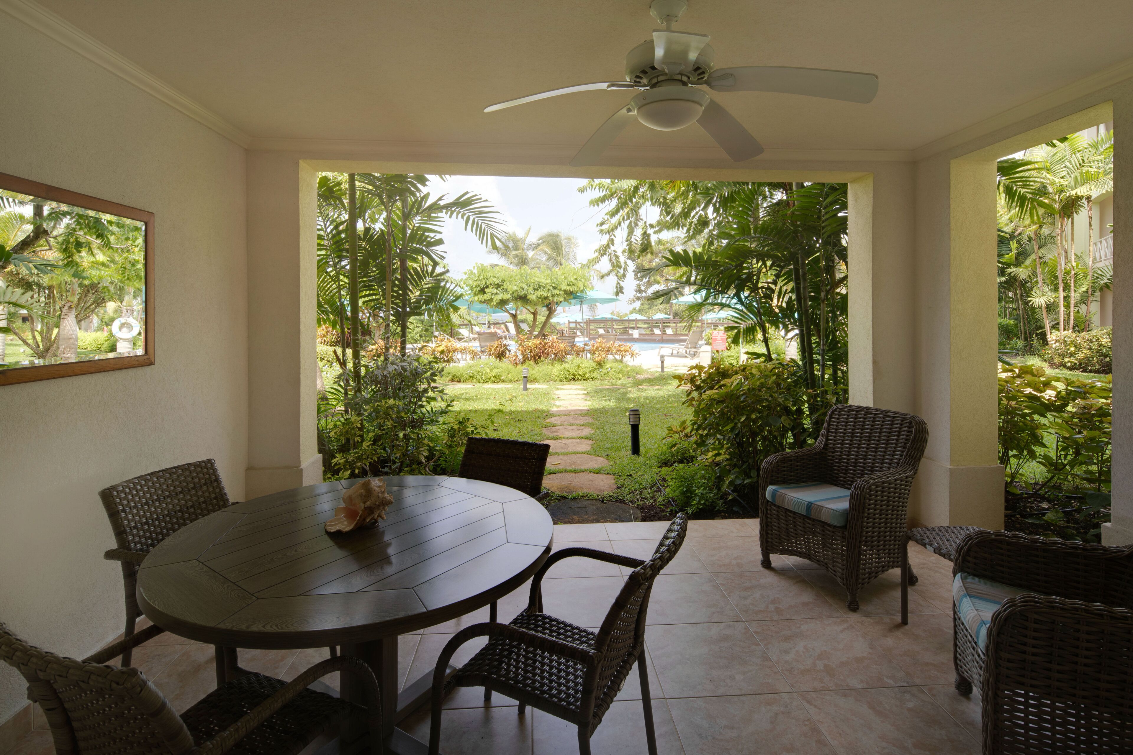 A patio with a round table and wicker chairs, a ceiling fan, and a view of a pool and garden.