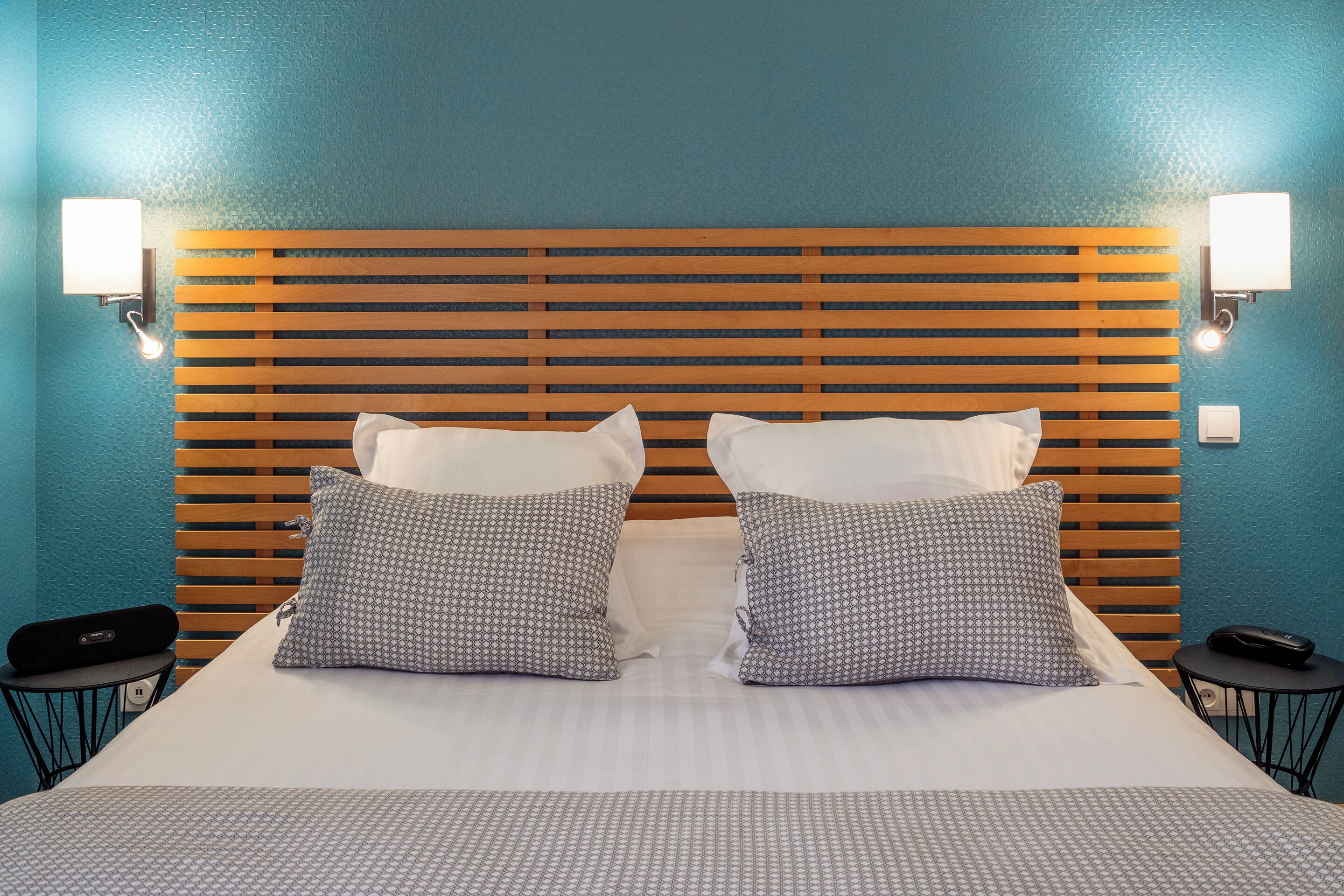 A hotel room with a wooden headboard, two patterned pillows, a white bedspread, and two bedside tables with lamps.