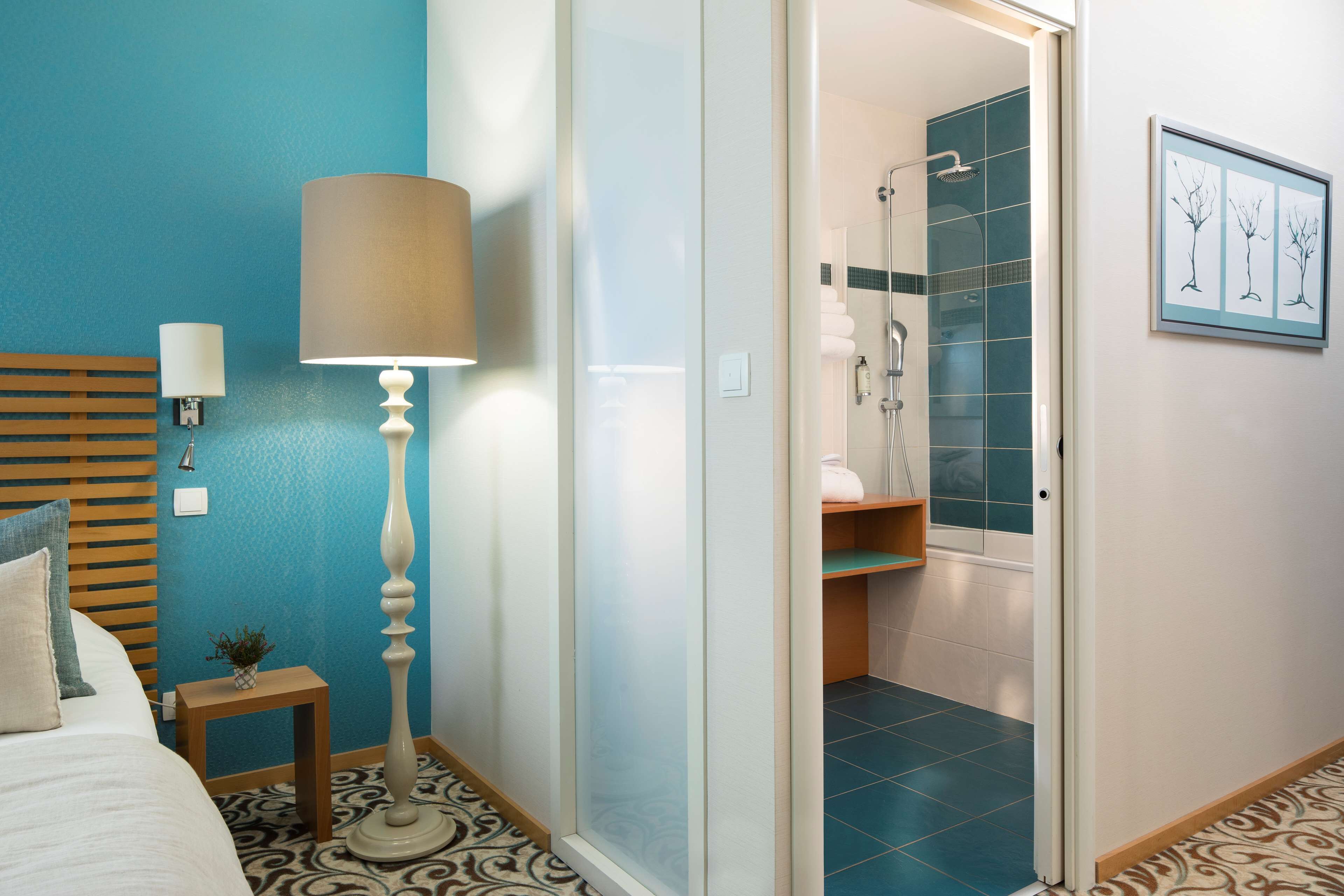 Une chambre d’hôtel comprenant un lit, une lampe de chevet, une table de chevet avec une plante, une grande lampe sur pied, une salle de bain avec douche et un tableau accroché au mur.