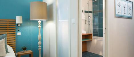 Habitación de hotel con cama, lámpara de noche, mesita de noche con una planta, una lámpara de pie grande, un baño con ducha y un cuadro en la pared.