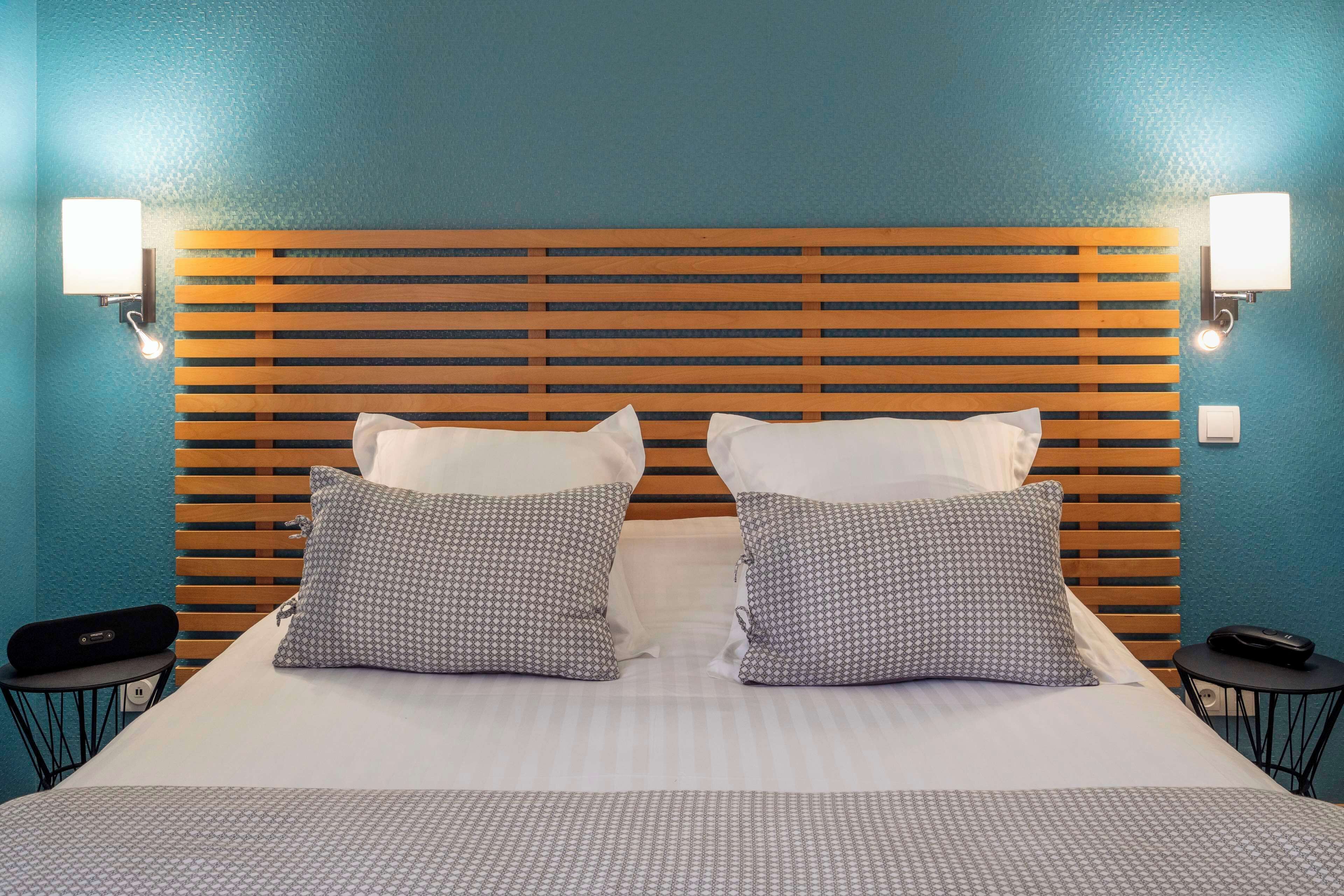 Quarto de hotel com cabeceira de madeira, duas almofadas estampadas, colcha branca e duas mesas de cabeceira com abajures.