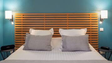 Quarto de hotel com cabeceira de madeira, duas almofadas estampadas, colcha branca e duas mesas de cabeceira com abajures.