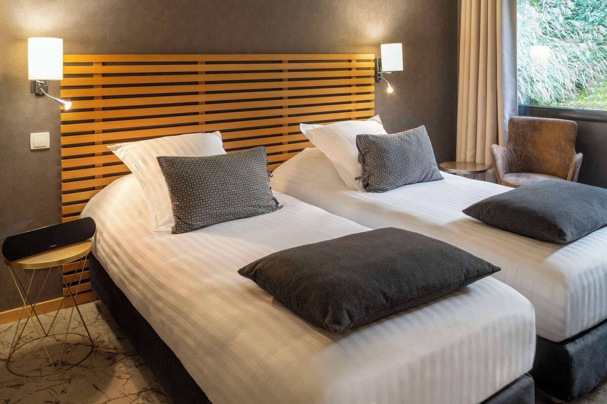 Een hotelkamer met twee bedden, een houten hoofdbord, bedlampen, een bijzettafeltje, een stoel en uitzicht op groen.