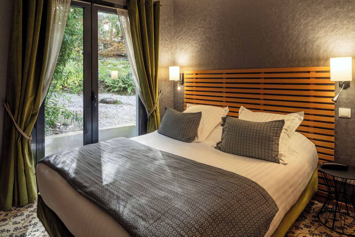 Een hotelkamer met een groot bed, een houten hoofdbord, twee bedlampen en uitzicht op groen buiten het raam.