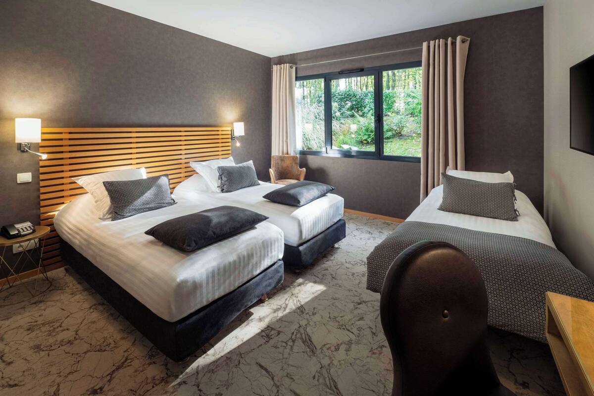 Een hotelkamer met twee bedden, een houten hoofdbord, een tafeltje, een stoel en een raam met gordijnen.