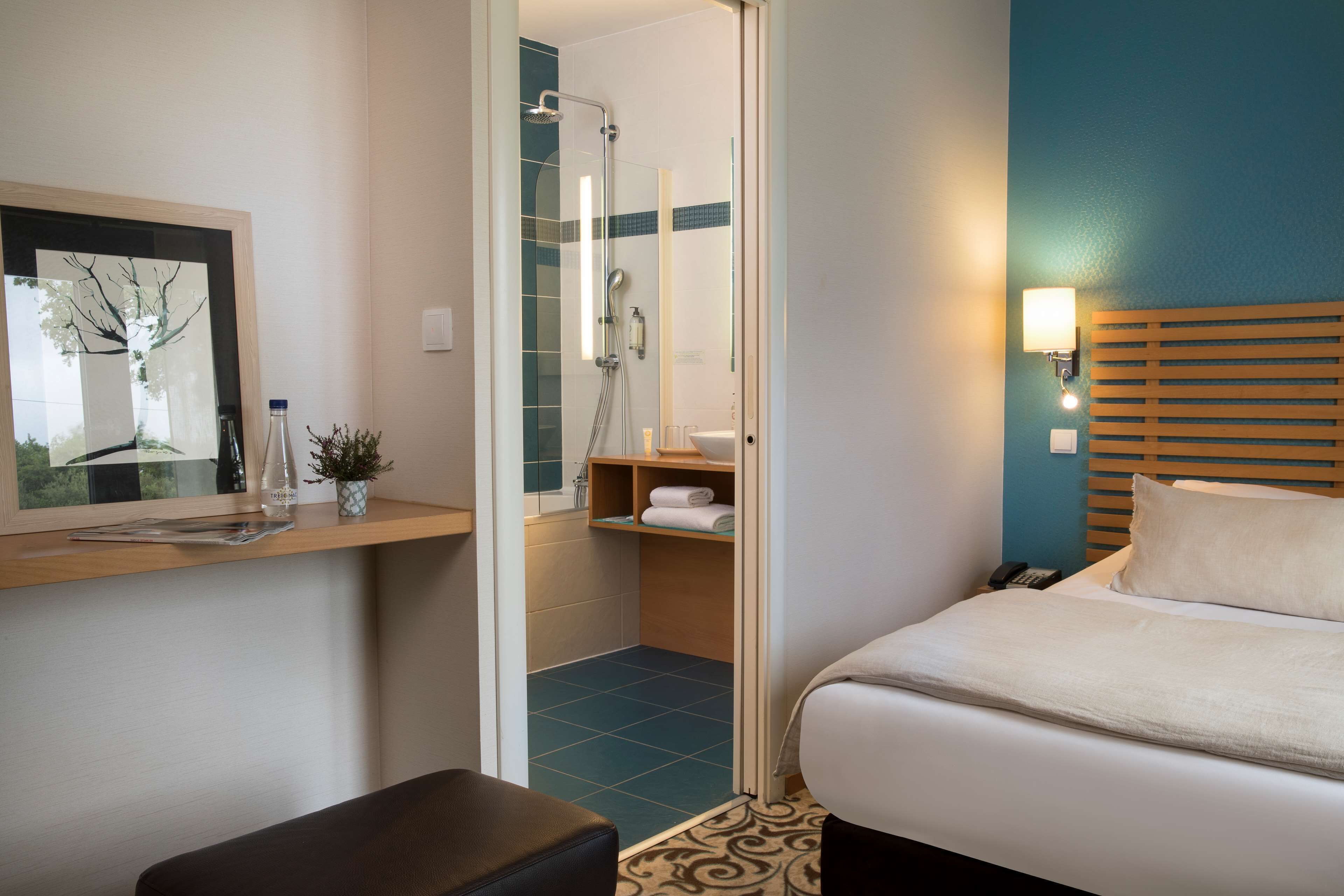 Habitación de hotel con cama, zona de ducha, una mesita con una planta y una lámpara de pared.