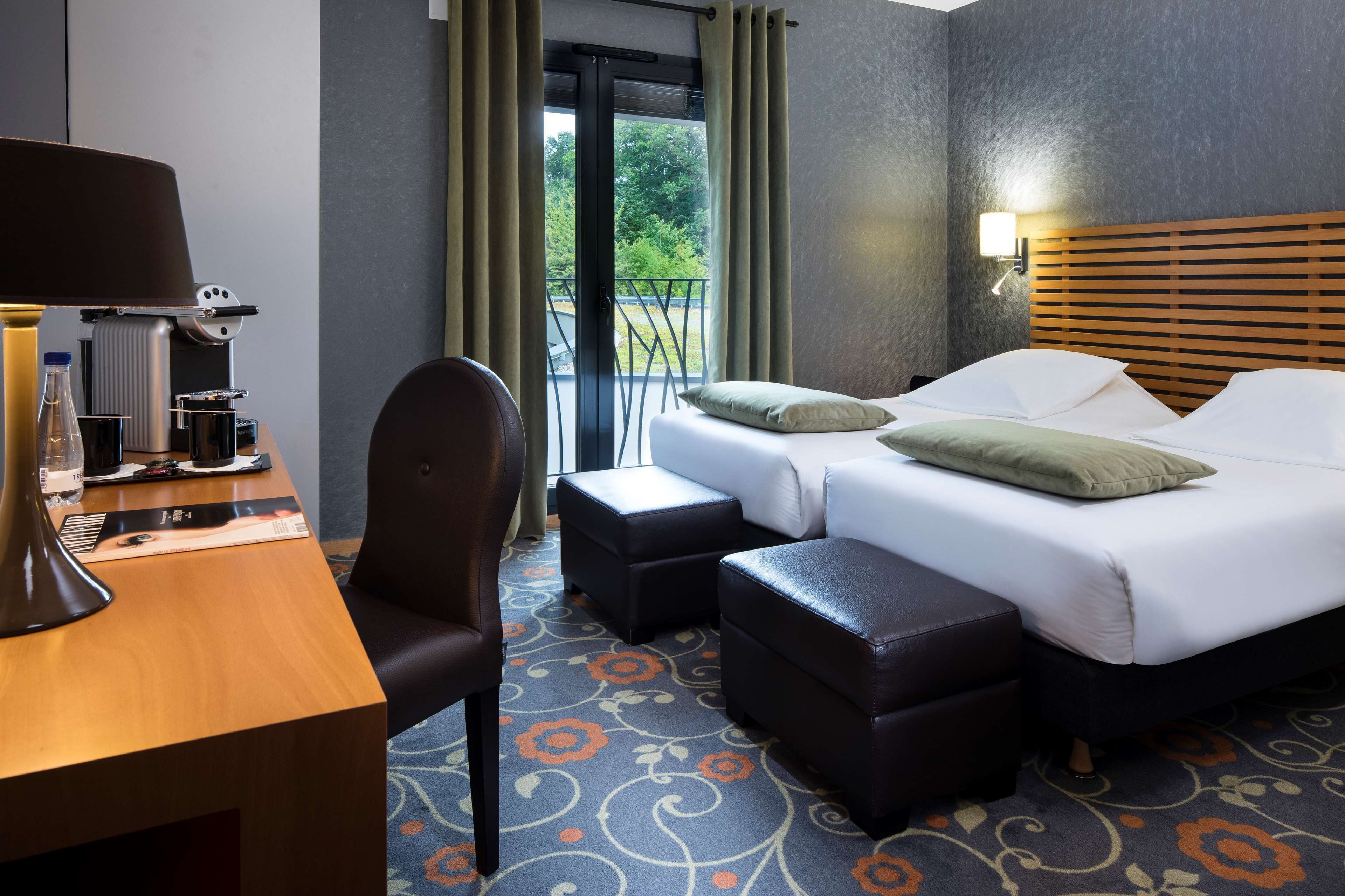 A hotel room with a desk, two beds, a chair, and a balcony with curtains.