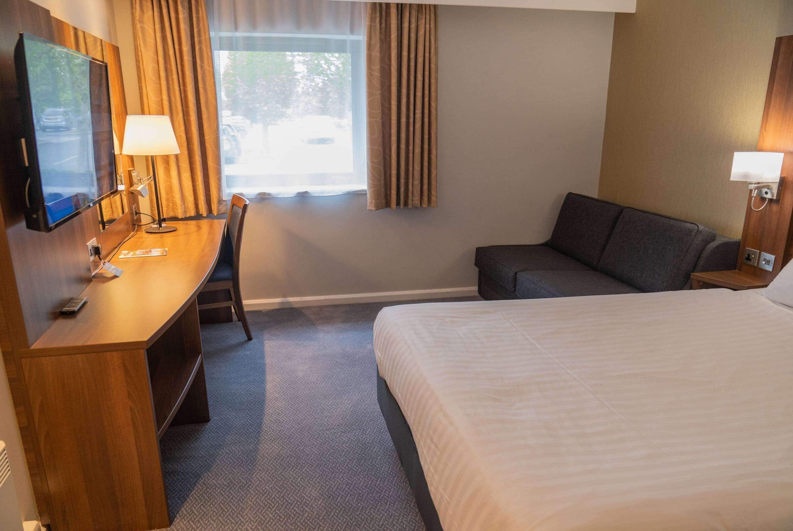 Habitación de hotel con cama, escritorio con televisor, sillón, sofá y ventana con cortinas.