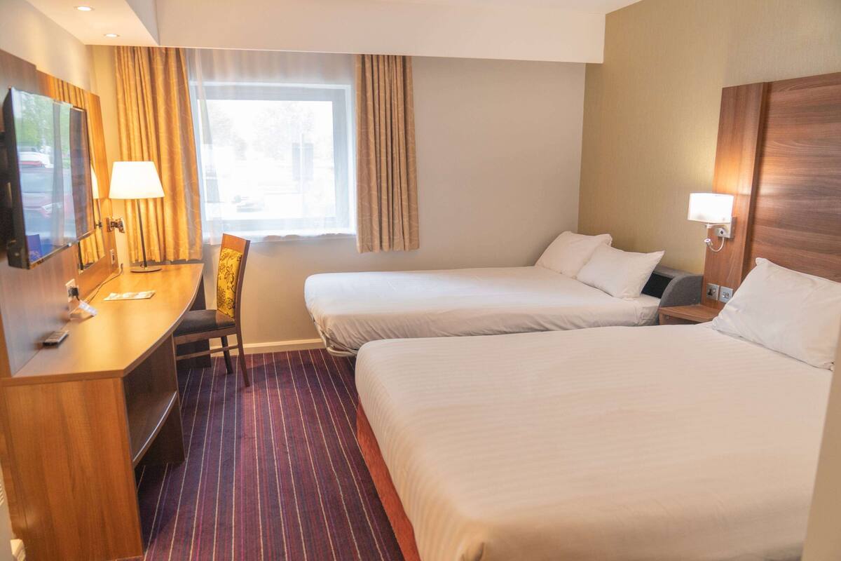 A hotel room with two beds, a desk with a chair, a TV, and a window with curtains.