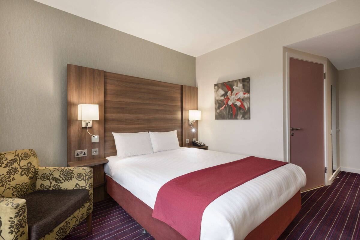 A hotel room with a large bed, a wooden headboard, two bedside lamps, a floral armchair, and a door to the right.